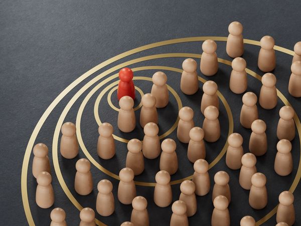 A red pawn stands out among many beige pawns on a black surface with golden concentric circles.