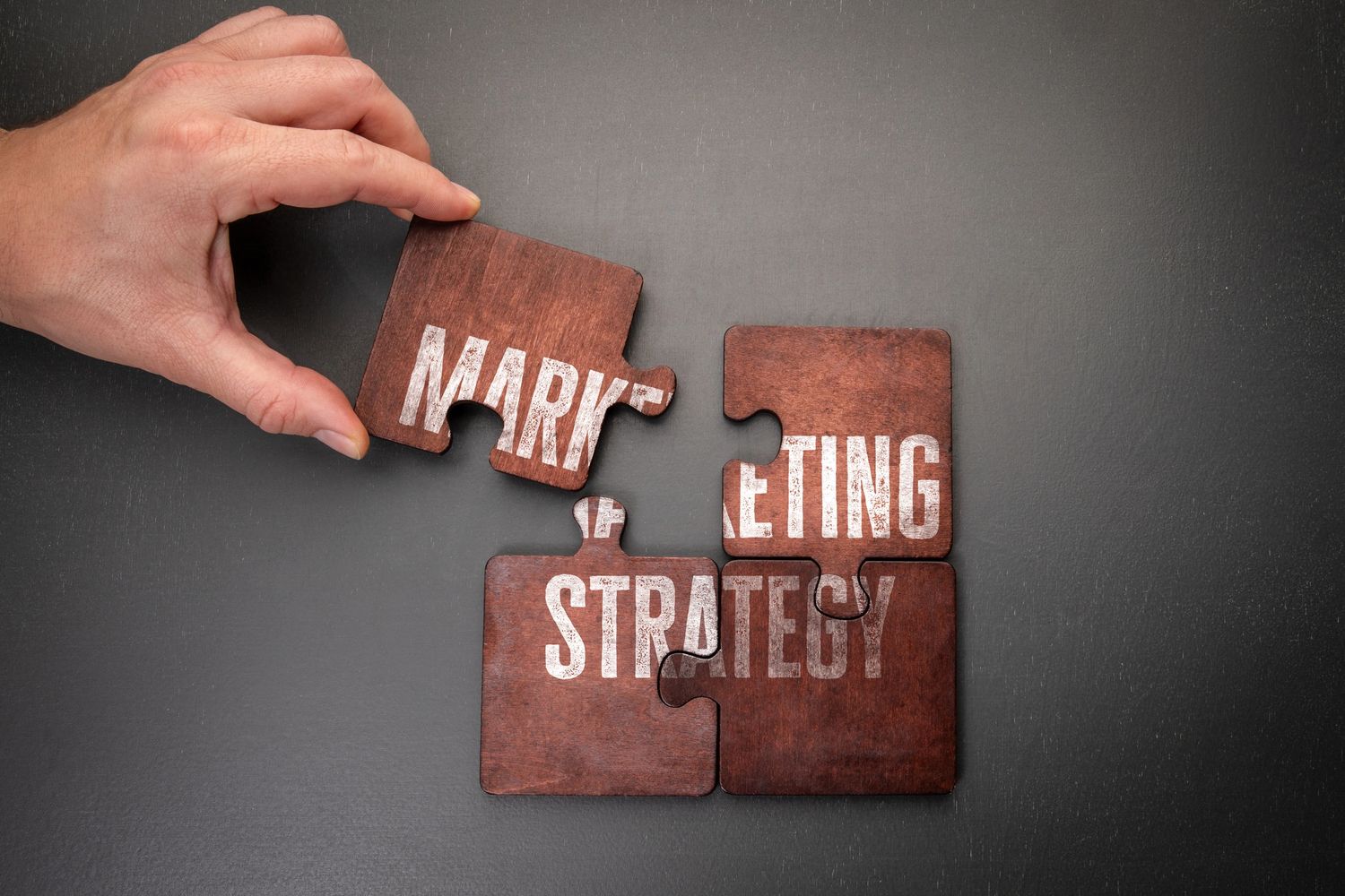 Hand placing a puzzle piece labeled 'MARK' to complete 'MARKETING STRATEGY'.