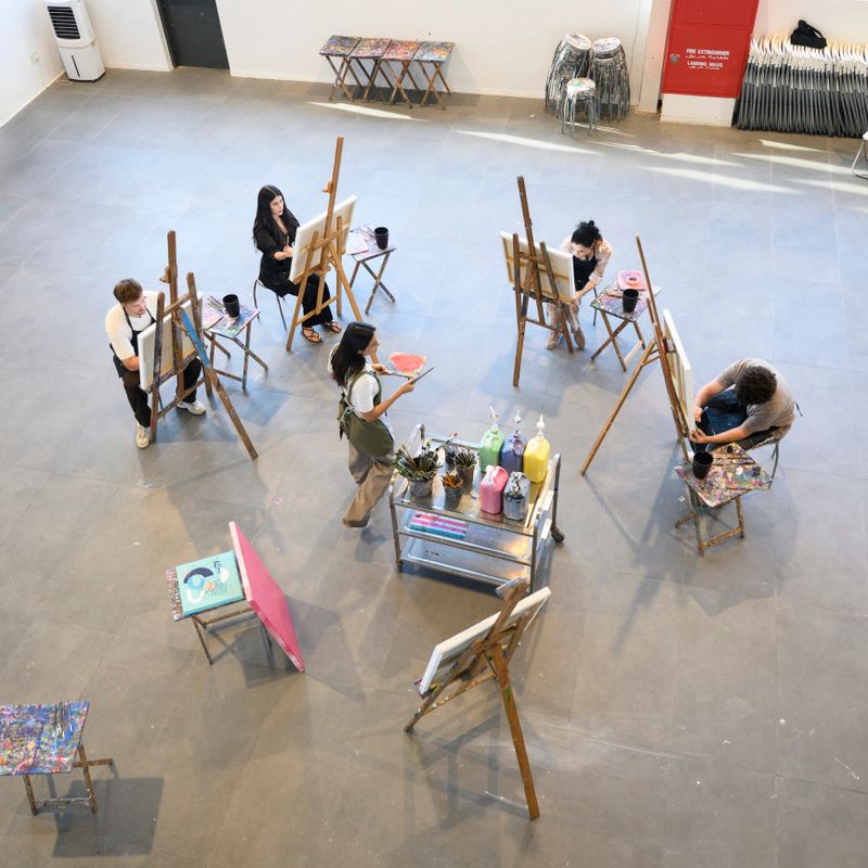 Elevated view of mature students and teacher in bright daylight art studio studying the art of painting on canvas. Easels and artists form a semi-circle around mid adult female teacher demonstrating with palette and paint.