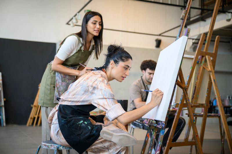 A young woman adult student is drawing on artists canvas in preparation for painting with guidance and advice from her female mentor and teacher. They are in a bright artists studio class.