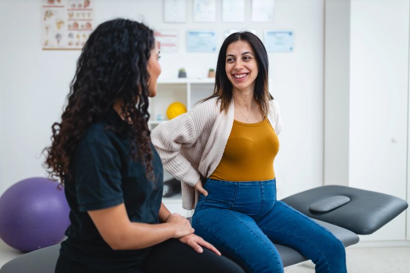 A Hispanic physiotherapist provides guidance to a pregnant woman in a medical clinic. They discuss healthcare strategies focused on pain relief and physical therapy for a healthy lifestyle.