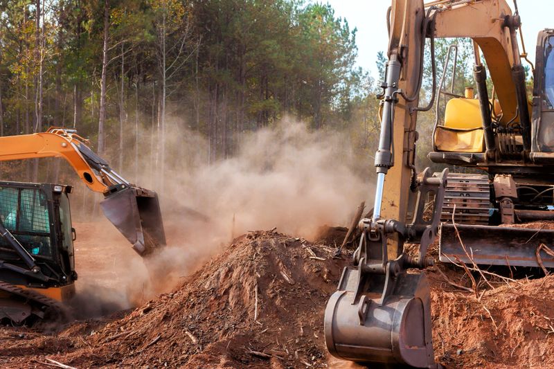 Bulldozers, excavators work together to clear trees, debris in forested area under at construction site