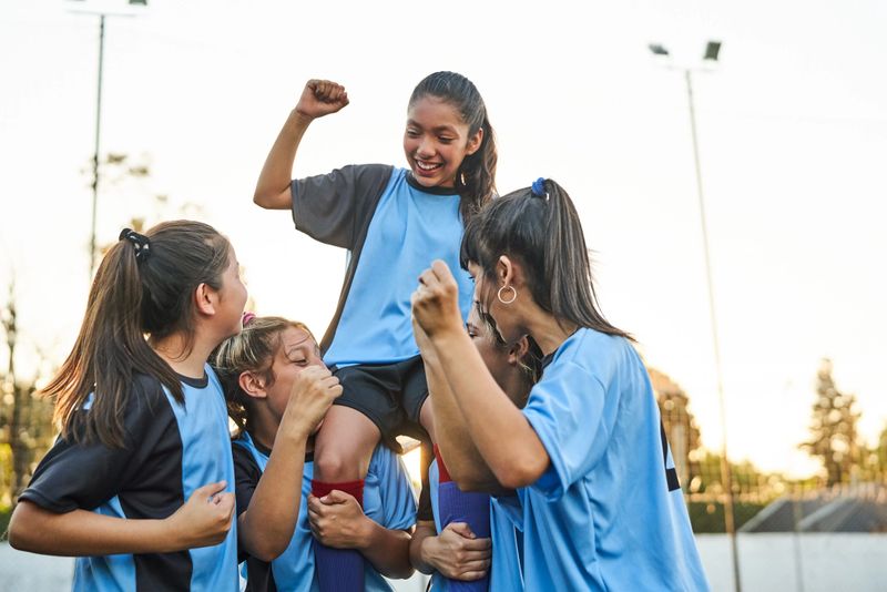 Female soccer team celebrating victory on the outdoor field, with players carrying a teammate on their shoulders. The team's joy and excitement highlight their triumph and unity.