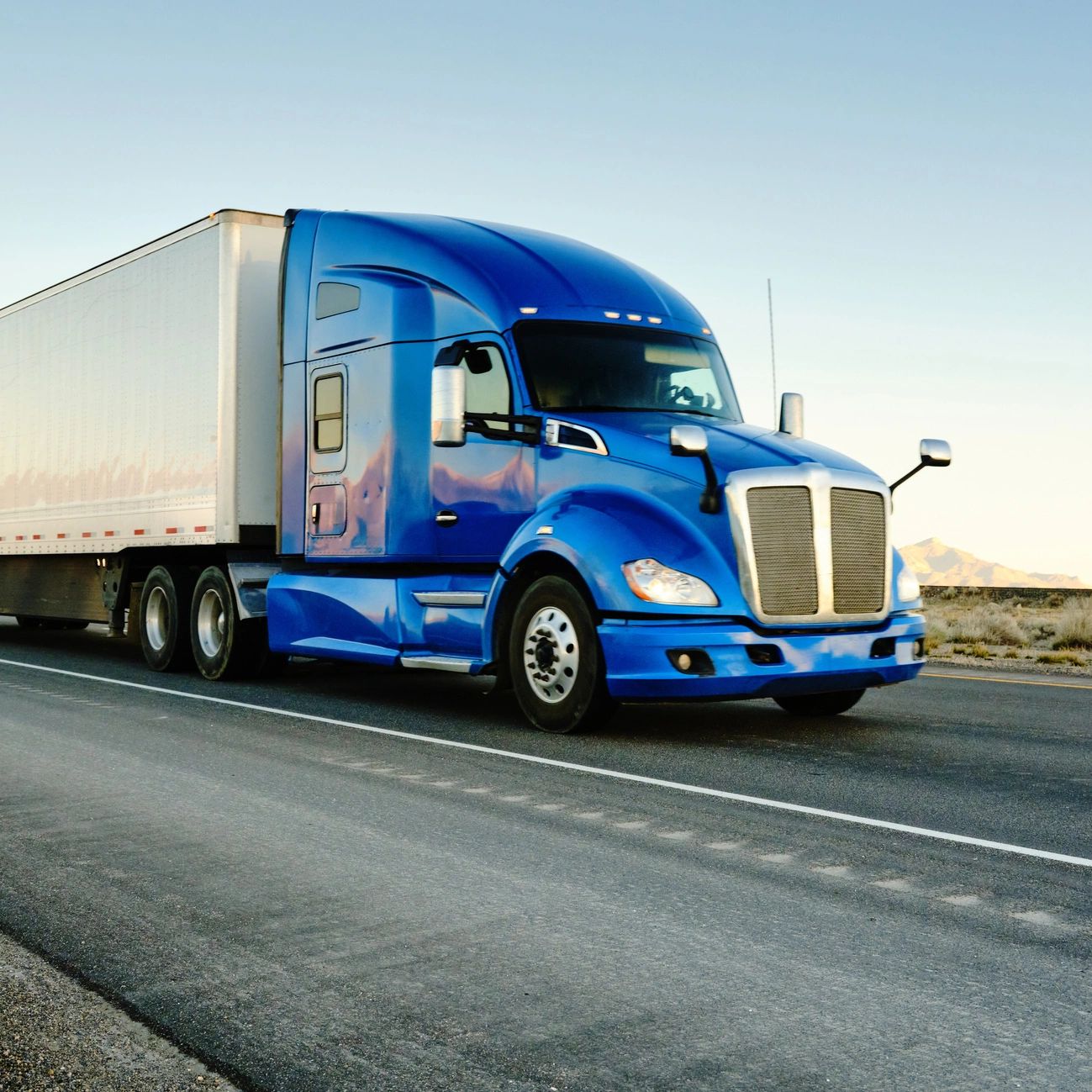 Blue semi-truck driving on an empty highway during sunset.