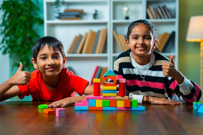 Happy Indian Asian Curious Little Girl and Boy Siblings Joyfully Playing with Colorful Wooden Block Toys, Building Structures, and Exploring Creativity Together While Sitting at Table in a Modern Home
