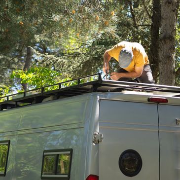 Man installing roof rack on a camper van in a forested area.