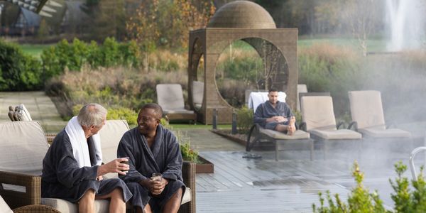 Two men in robes chatting by a spa pool with another man relaxing in the background.