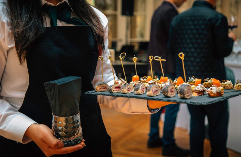 A professional waitress in a white shirt and black apron presenting a selection of elegantly prepared appetizers on a slate tray. The skewered bites include a variety of gourmet options, perfect for formal gatherings or high-end events. The focus is on the tray and its details, highlighting the sophistication of the cuisine and the attentiveness of the service
