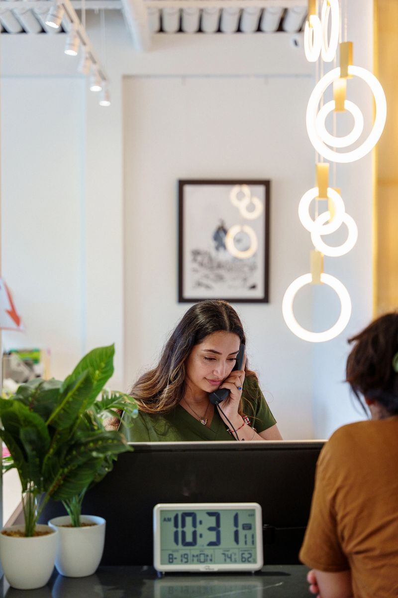 A female receptionist of Hispanic descent working at a dental clinic talks with a patient on the telephone while a patient waits at the reception desk to be checked in for their appointment.