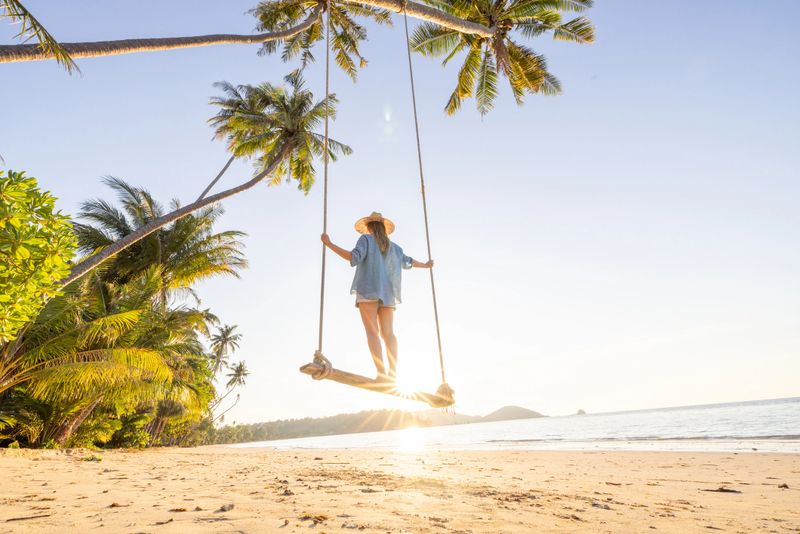 Woman in sun hat enjoying beach swing at sunset. Peaceful tropical coastline with palm trees. Vacation idyll on a serene sandy beach.