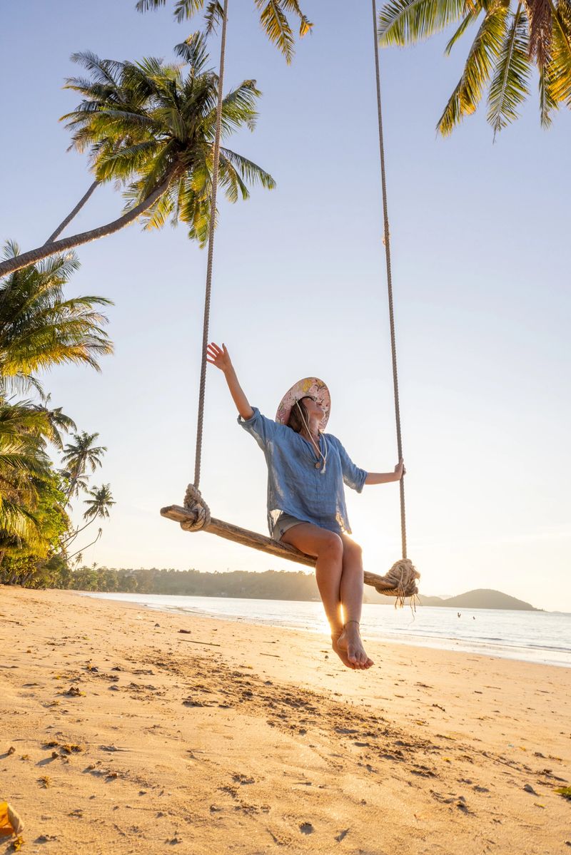 Woman in a sun hat enjoying a swing on a tropical beach at sunset. Relaxed beach scene with palm trees.