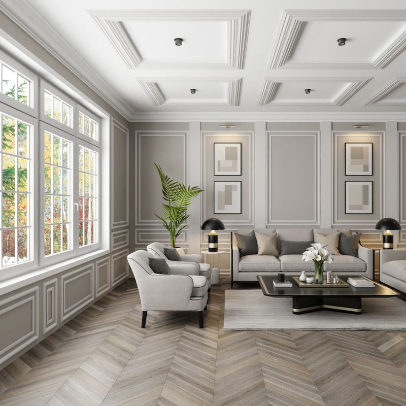 Experience the charm of modern neo-classical Parisian interior design with this luxurious living room setup. The space features classic wall molding plaster design and large classic windows that flood the room with natural light. The sophisticated beige color palette is complemented by plush pillows and sleek modern furniture. A stylish living room table with golden accents and a striking vase filled with white flowers add the finishing touches. This image is ideal for showcasing modern furniture, elegant decor, clean design, cozy room ideas, and renovation projects with a modern classic style.