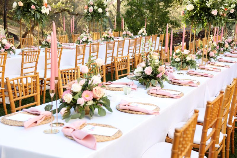 This beautifully styled outdoor wedding table features delicate floral centerpieces of pastel roses and greenery, paired with soft pink taper candles for a romantic glow. The setup includes white tablecloths, natural wooden chargers, and pink napkins, all surrounded by elegant wooden chairs. A serene and picturesque setting for a memorable wedding reception.