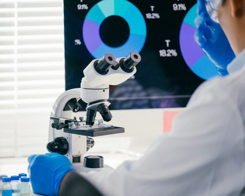 Scientist wearing gloves conducts research, analyzing samples under a microscope in a laboratory setting with a computer displaying data charts in the background