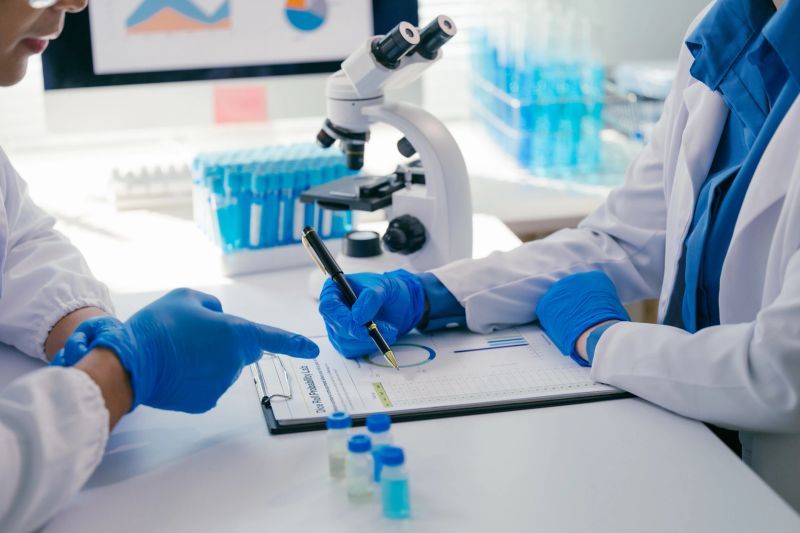 Two scientists wearing lab coats and gloves are examining data on a clipboard, discussing research findings in a modern laboratory setting with a microscope and test tubes