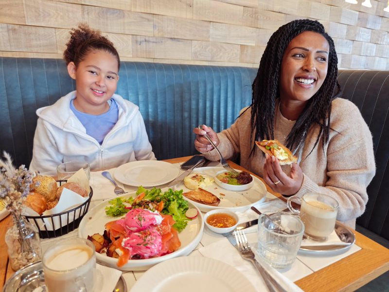 A cheerful mother and her daughter enjoy a vibrant meal together at a cozy café. The table is adorned with colorful dishes, fresh bread, and creamy lattes, creating a warm and inviting atmosphere. Their smiles and connection highlight a moment of happiness and togetherness.