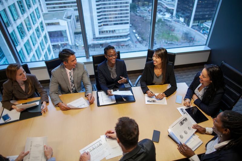 High angle view of Black ethnicity mid adult woman businesswoman with multiracial group of work colleagues in business meeting talking discussing plans ideas in businesswear in board room conference table with urban skyline cityscape