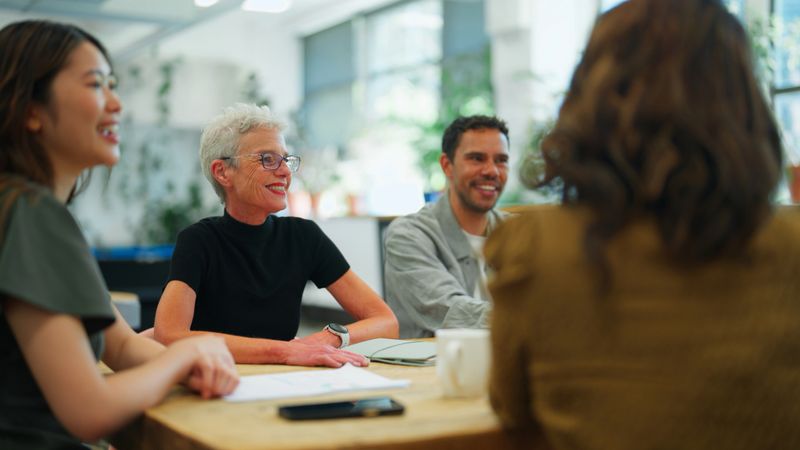 A diverse group of businessmen and businesswomen engage in a collaborative discussion around a large wooden table. A laptop is open as part of the conversation, emphasizing the use of technology in teamwork. The office is bright, filled with natural light and greenery, reflecting sustainability and a modern, inclusive workspace culture. Senior adult, mature adult, young adult, Asian ethnicity, Southeast Asian ethnicity, multi-racial person, Aboriginal ethnic man and transgender businesswomen and businessman are working together.