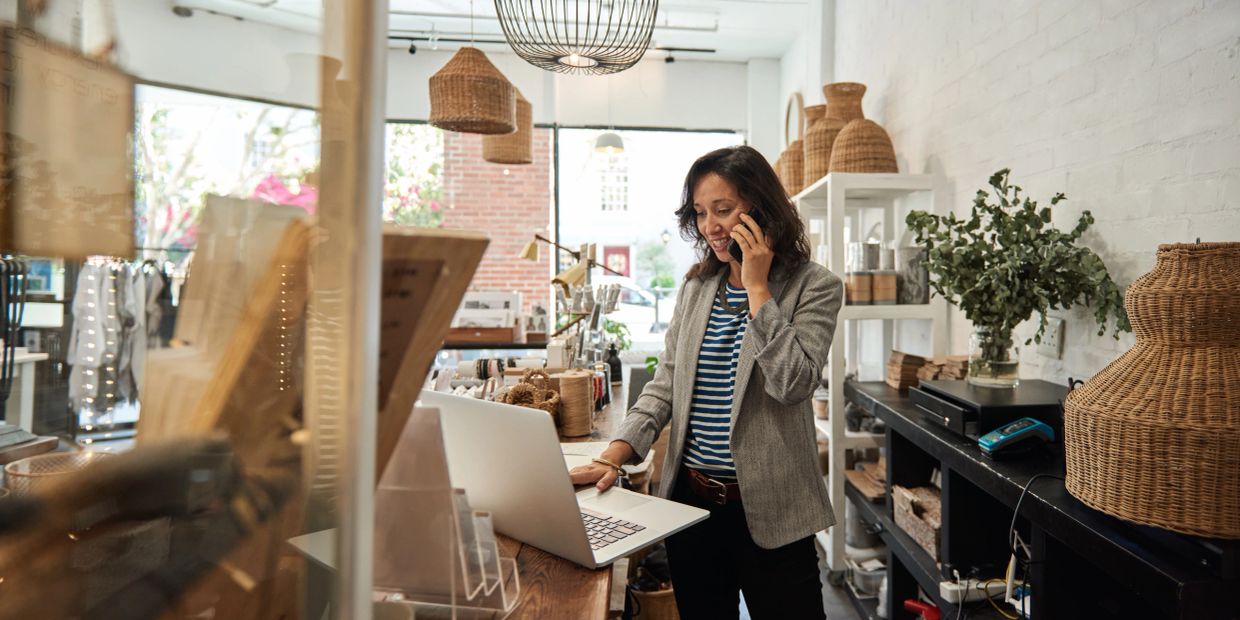 Woman talking on phone using laptop in a cozy store.