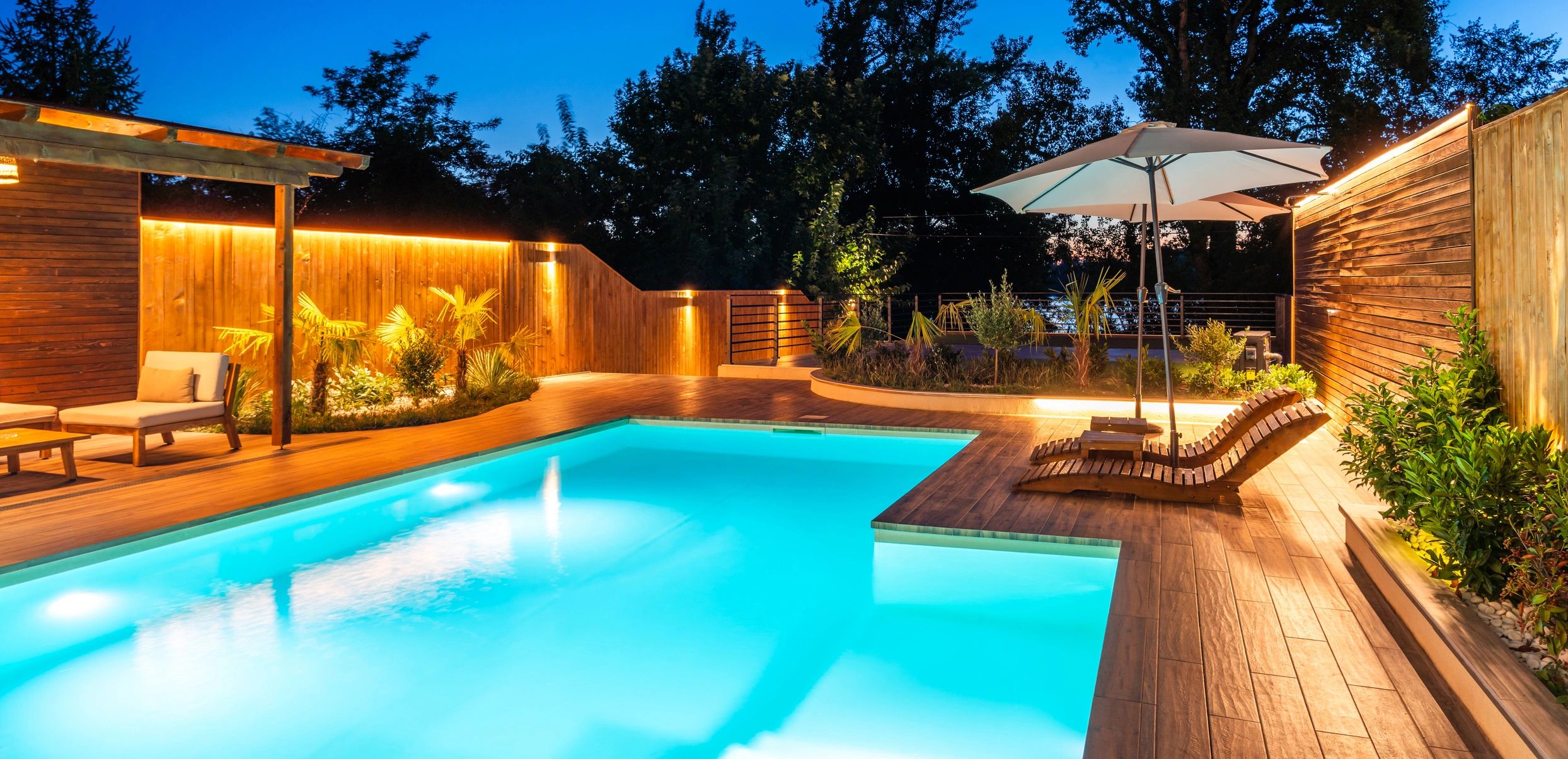 Modern backyard with illuminated pool and wooden deck at dusk.