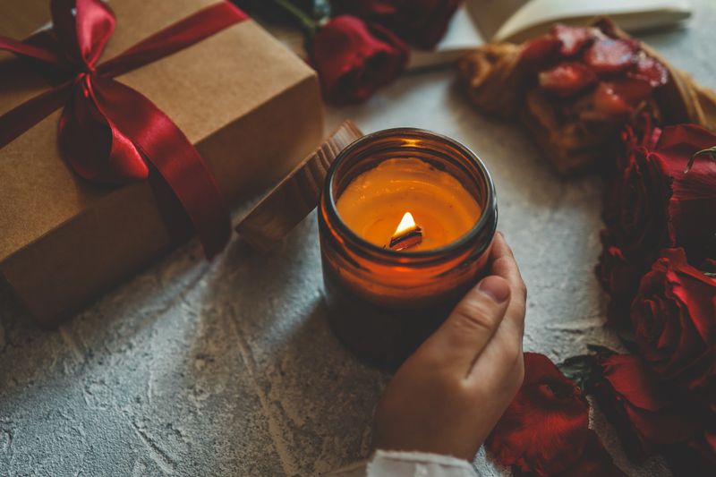 A glowing amber jar candle with a wooden lid, placed next to a kraft gift box tied with a red ribbon. Surrounded by fresh red roses, petals, and a pastry, creating a romantic, cozy ambiance.