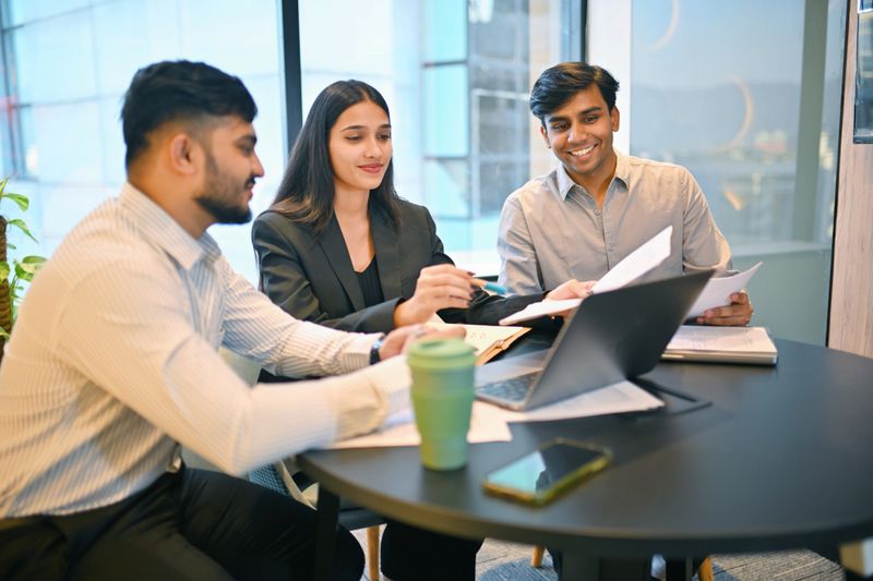 A group of young business professionals engaged in a dynamic discussion around a table in a contemporary meeting room. The atmosphere is energetic and collaborative, with laptops, notebooks, and coffee cups spread across the table. Large windows allow natural light to flood the space, highlighting the team's focus and creativity as they share ideas and strategize together
