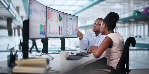 Two colleagues analyzing data charts on dual monitors at an office desk.