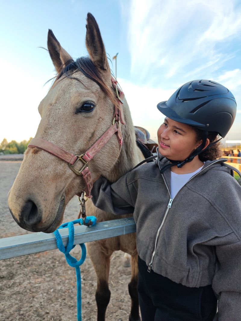 A young girl wearing a riding helmet stands beside her horse, gently resting her hand on the animal's neck. The serene stable setting highlights the strong bond between the girl and the horse, capturing a moment of connection and appreciation for nature and equestrian life.
