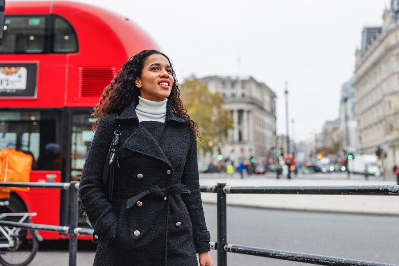 A cheerful Hispanic woman enjoys her solo adventure in London. She walks confidently, capturing the essence of exploration and joy. The iconic red double-decker bus in the background highlights her travel experience.