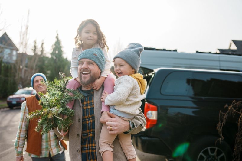 A handsome Caucasian man in his 30s who is carrying a a Christmas tree into his house with his father-in-law's help, holds his two young Eurasian kids as they laugh together.