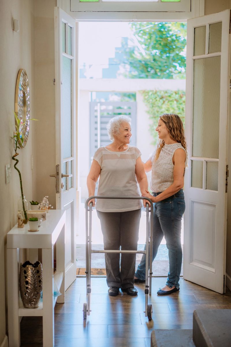 An elderly woman using a walker is assisted by a smiling young woman in a bright, welcoming home setting, illustrating care, support, and intergenerational bonding.