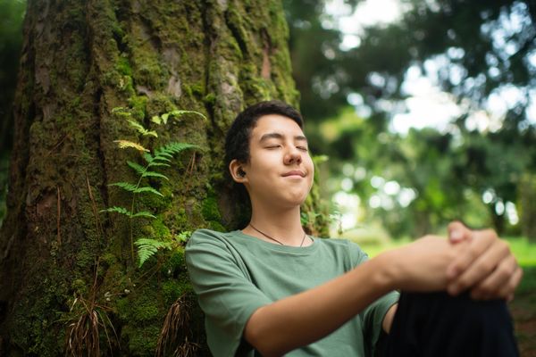 Young person meditating peacefully against a moss-covered tree in nature.
