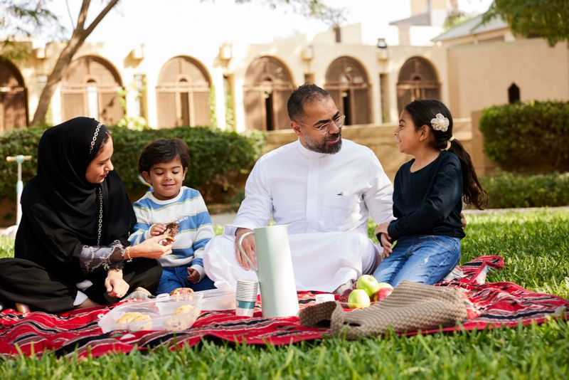 Sitting on the grass, a family with two children shares a picnic in a Riyadh park on a sunny weekend.