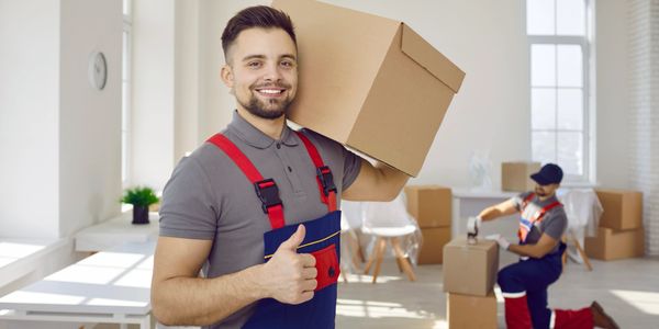 Smiling mover holding a box and giving a thumbs up in a bright room.