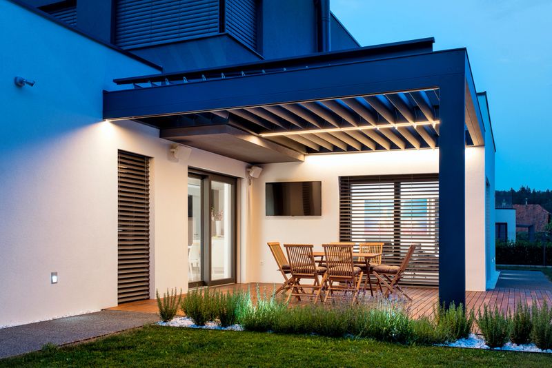 A modern patio with wooden furniture, featuring a table and chairs under a stylish pergola. The area is well-lit, showcasing a contemporary design with sliding glass doors and a neatly landscaped lawn.
