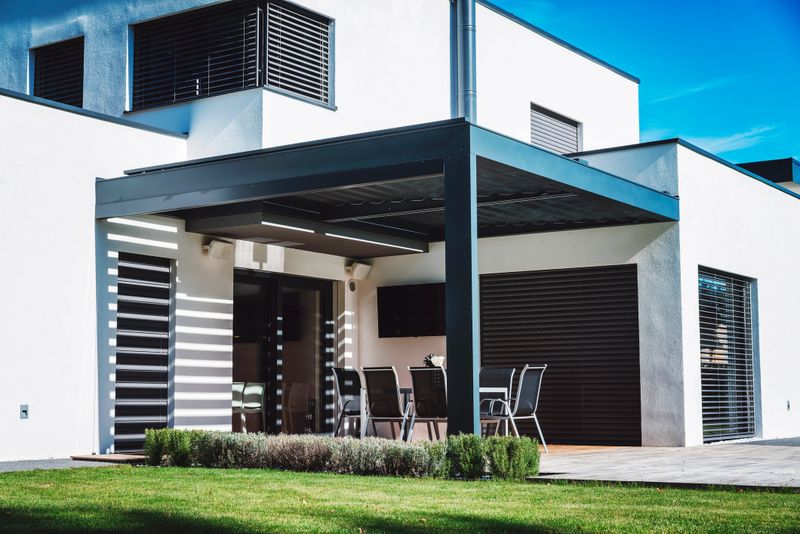 A modern house exterior featuring a patio with outdoor furniture, surrounded by green grass and shrubs. The design includes large windows and a sleek, minimalist style.