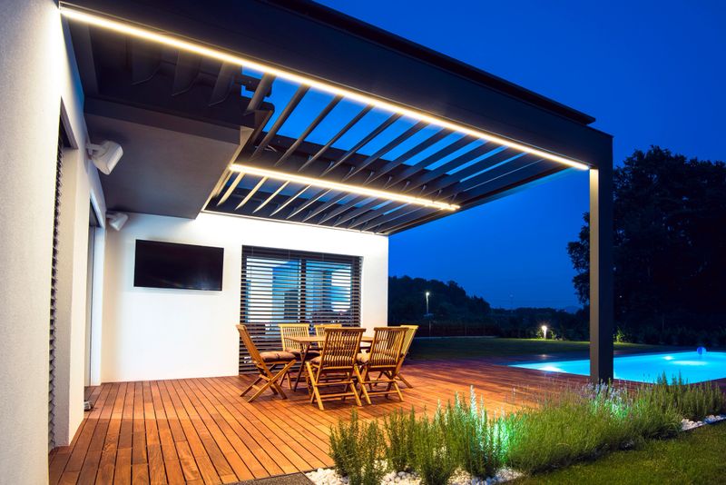 A modern outdoor patio with wooden furniture, illuminated by soft lighting. The setting features a swimming pool in the background and a sleek architectural design, creating a serene evening atmosphere.