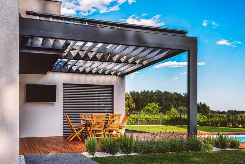 A modern outdoor patio with a pergola, featuring wooden furniture and a swimming pool in the background. The setting is surrounded by greenery and a clear blue sky.