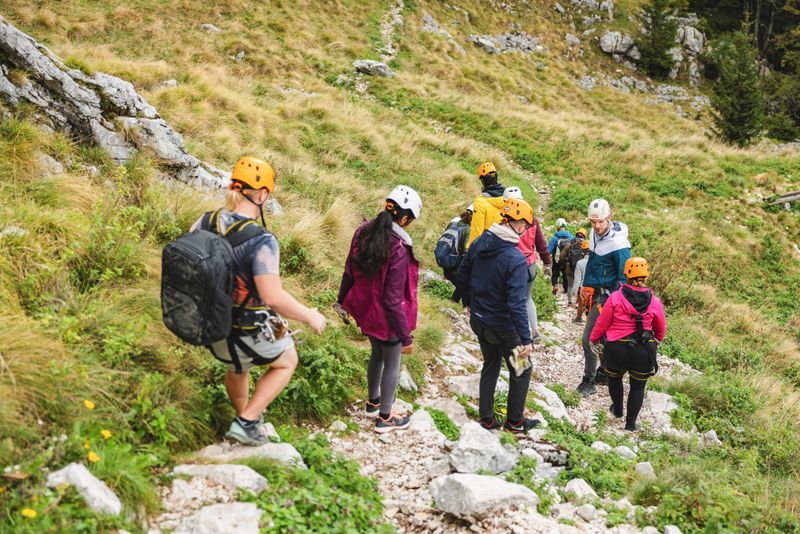 A diverse group of male and female friends equipped with helmets and harnesses hiking on a mountain trail, preparing for a zipline experience in a natural, green environment.