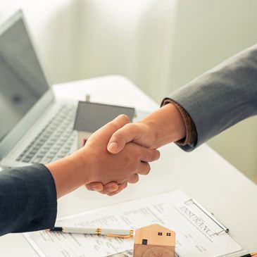 Two people shaking hands over a real estate deal with money and insurance documents.