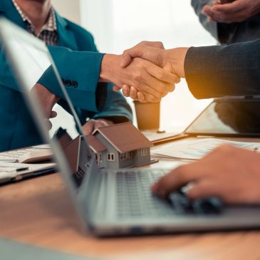 Two people shaking hands over a desk with a laptop and house models.