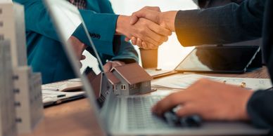 Two people shaking hands over a laptop with a small house model on the table.