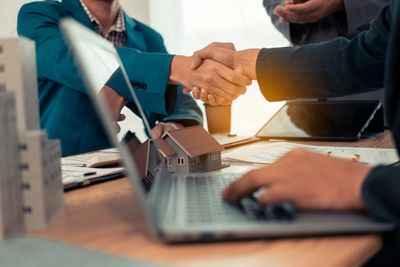 Two professionals shaking hands over a real estate deal with a laptop and house model.