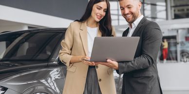Two professionals discuss something on a laptop near a car inside a showroom.