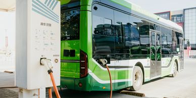 A green electric bus charging at a station under a canopy.