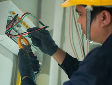 Electrician wearing gloves and a helmet using a multimeter to check wiring.