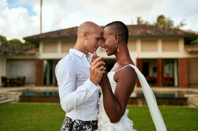 Ambiguous couple dances at tropical villa. Black person in white dress as bride partner in bold suit as LGBTQ groom. Wedding ceremony with love and joy. Inclusive celebration in exotic location.