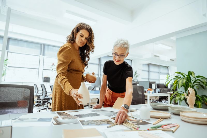 Two women collaborating at a brightly lit office desk reviewing documents and discussing creative ideas together