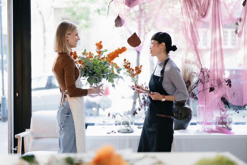 Two female florists working together in a flower shop, arranging floral decorations with a creative backdrop and stylish decor. The scene conveys a sense of creativity, teamwork, and small business operation.