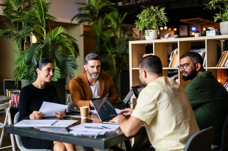 Cheerful businesswoman and three male co-workers sitting at table having conversation, teamwork, strategic planning, project briefing